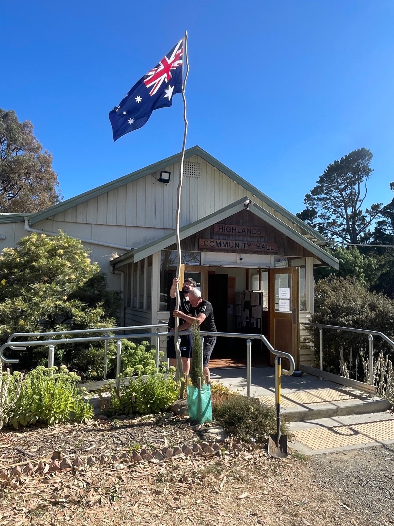 Two men raising the Australian flag on a bush pole outside the community hall