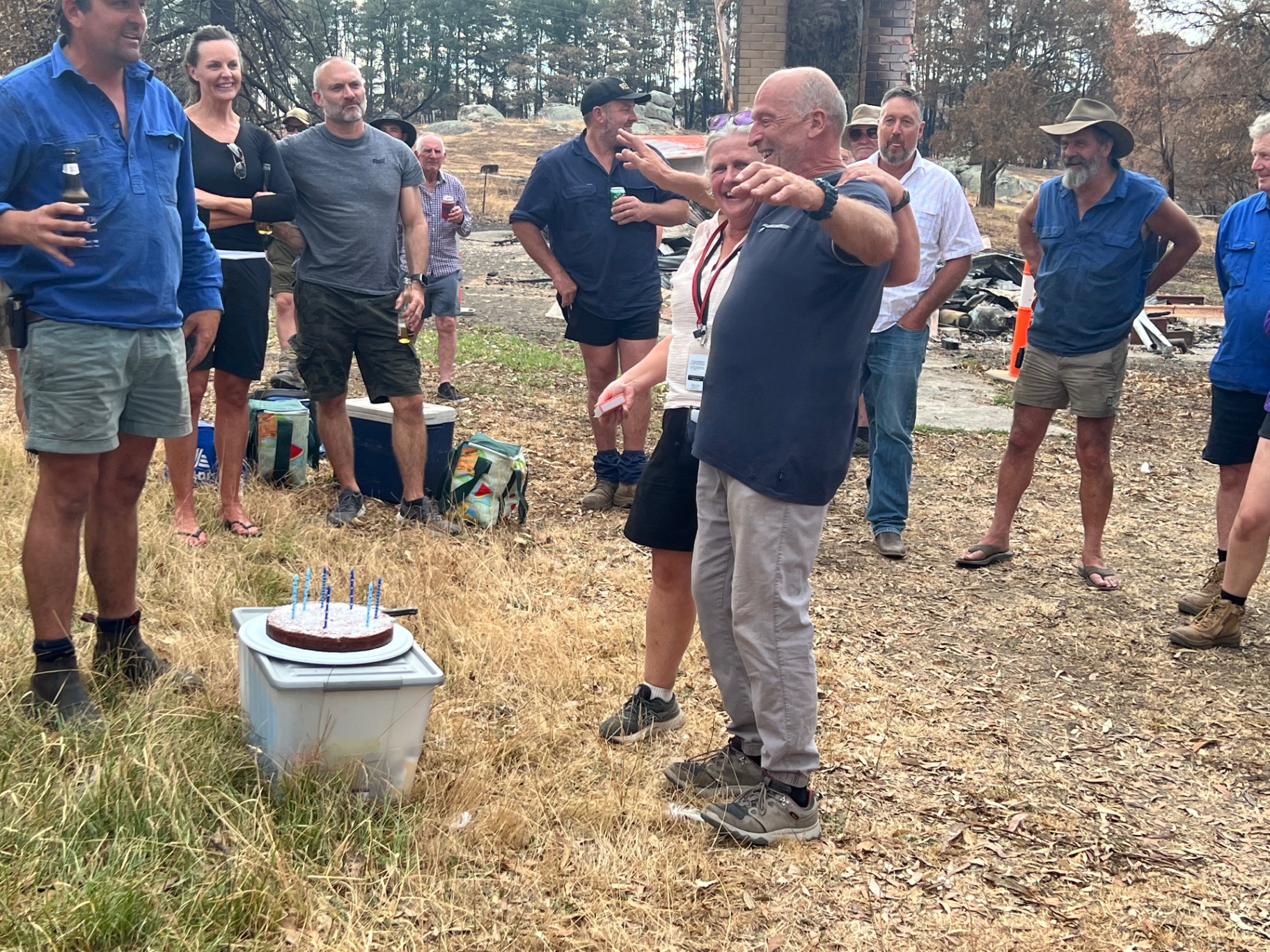 A group of locals gathered outdoors around a birthday cake on a rebuilt property